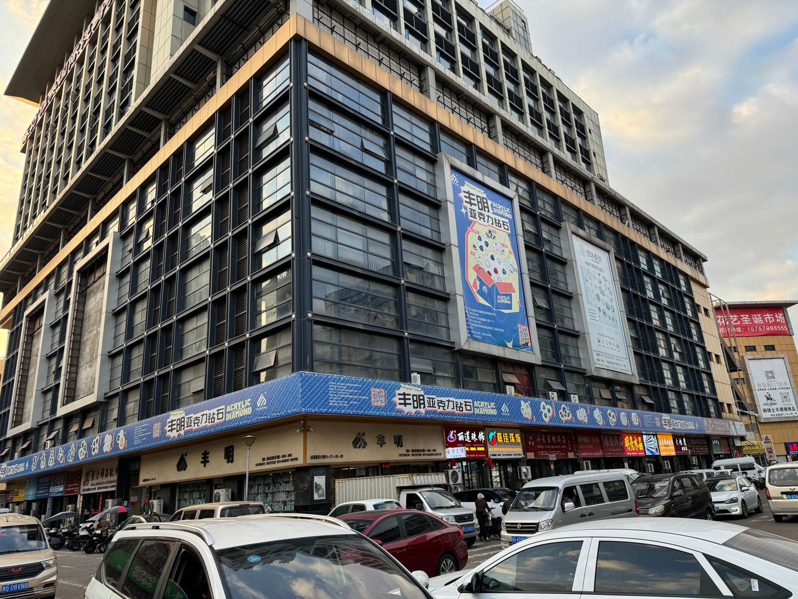Corner of Jinfuyuan Plaza in Yiwu, China, showing busy traffic and ground-floor jewelry-accessory stores advertising acrylic diamond rhinestones, cup chain, hot-fix, beads, chains and assorted DIY findings.