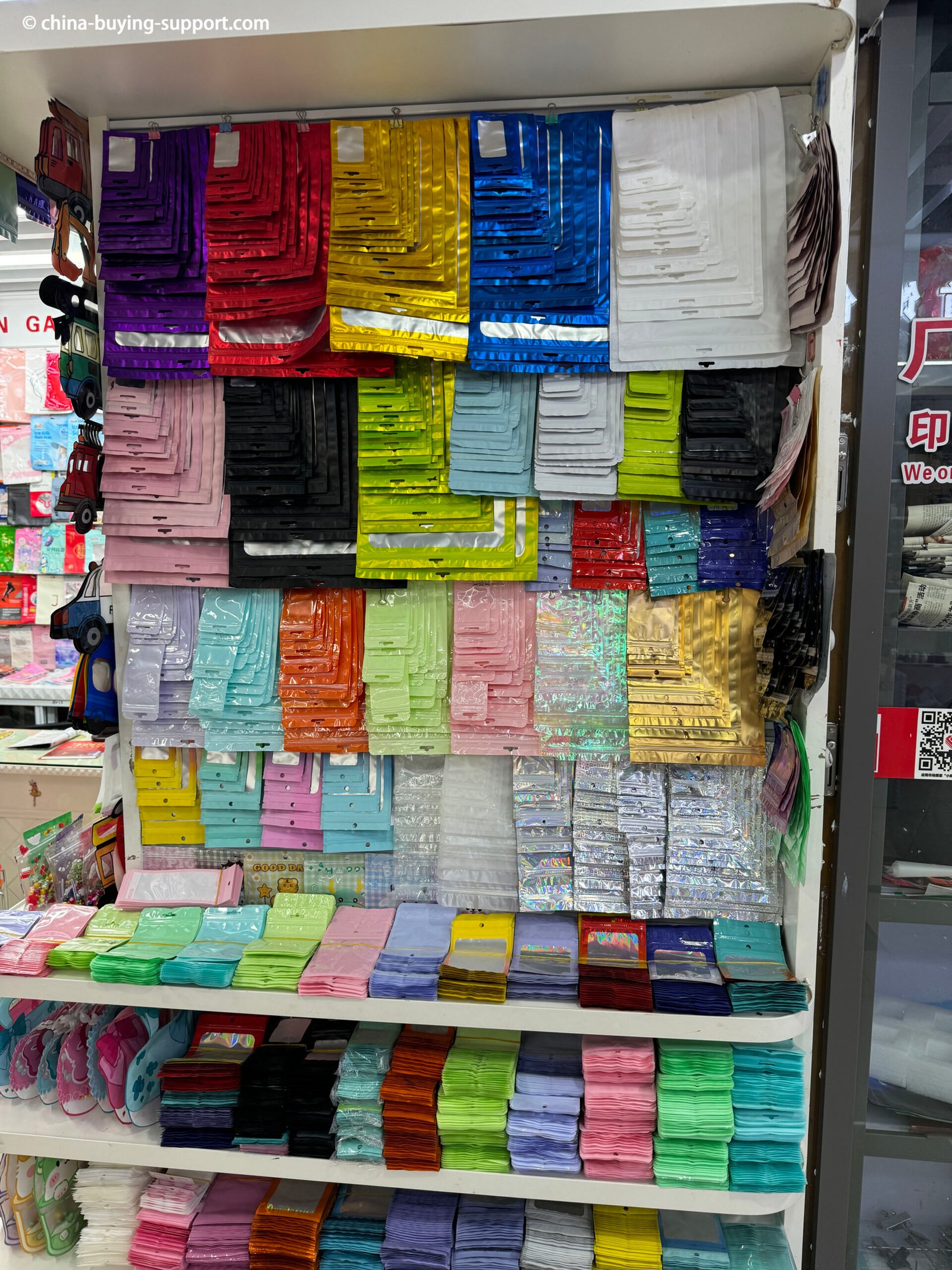 Rows of colorful metallic PVC packaging bags and jewelry pouches displayed on shelves at Yiwu International Trade City Gift Packaging Market, featuring glossy, holographic, and transparent finishes in various sizes and vibrant colors for jewelry and accessories.