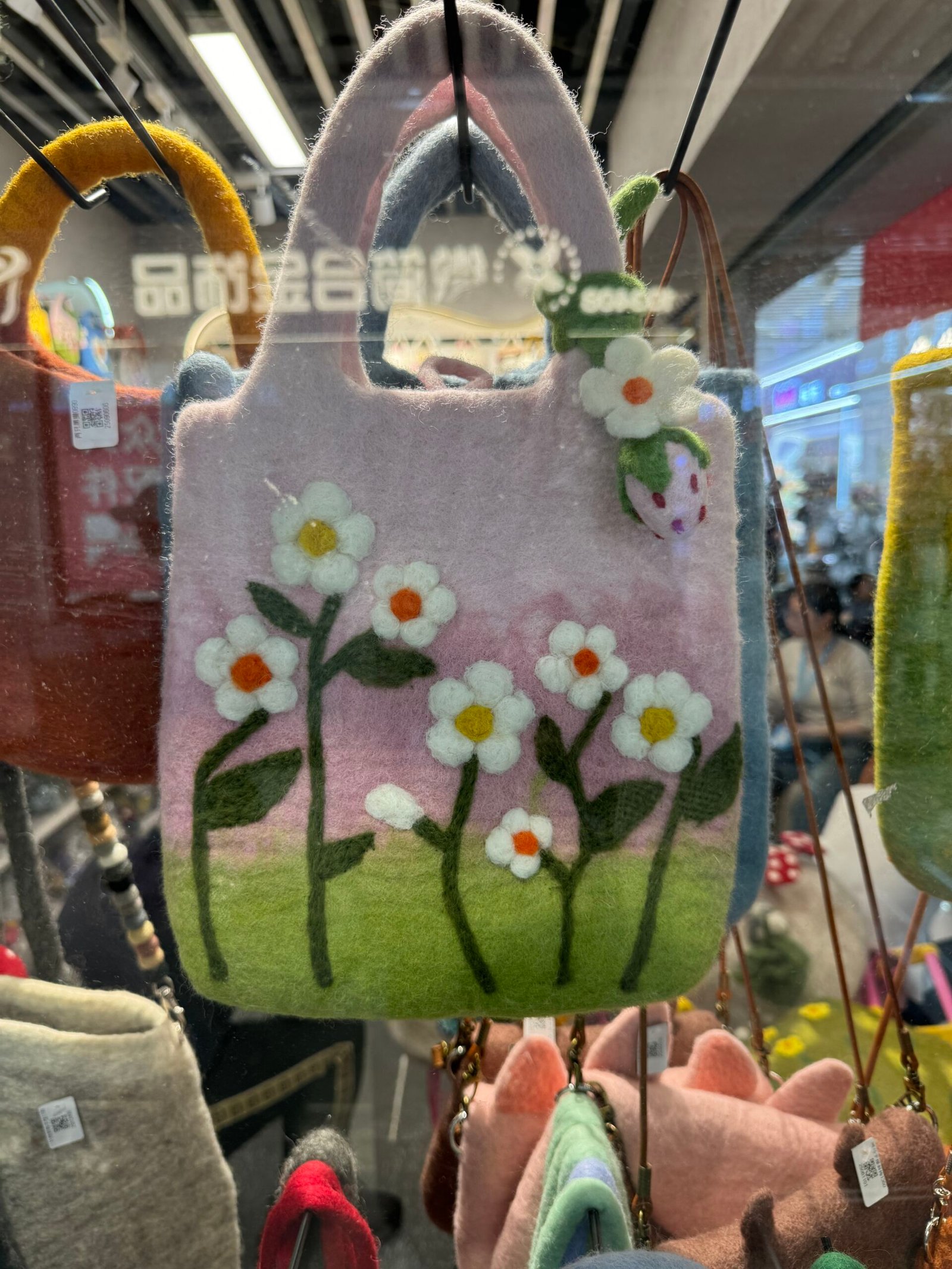 Close view of a needle-felted wool handbag with white daisy flowers and leaves, displayed in a Yiwu accessory shop.