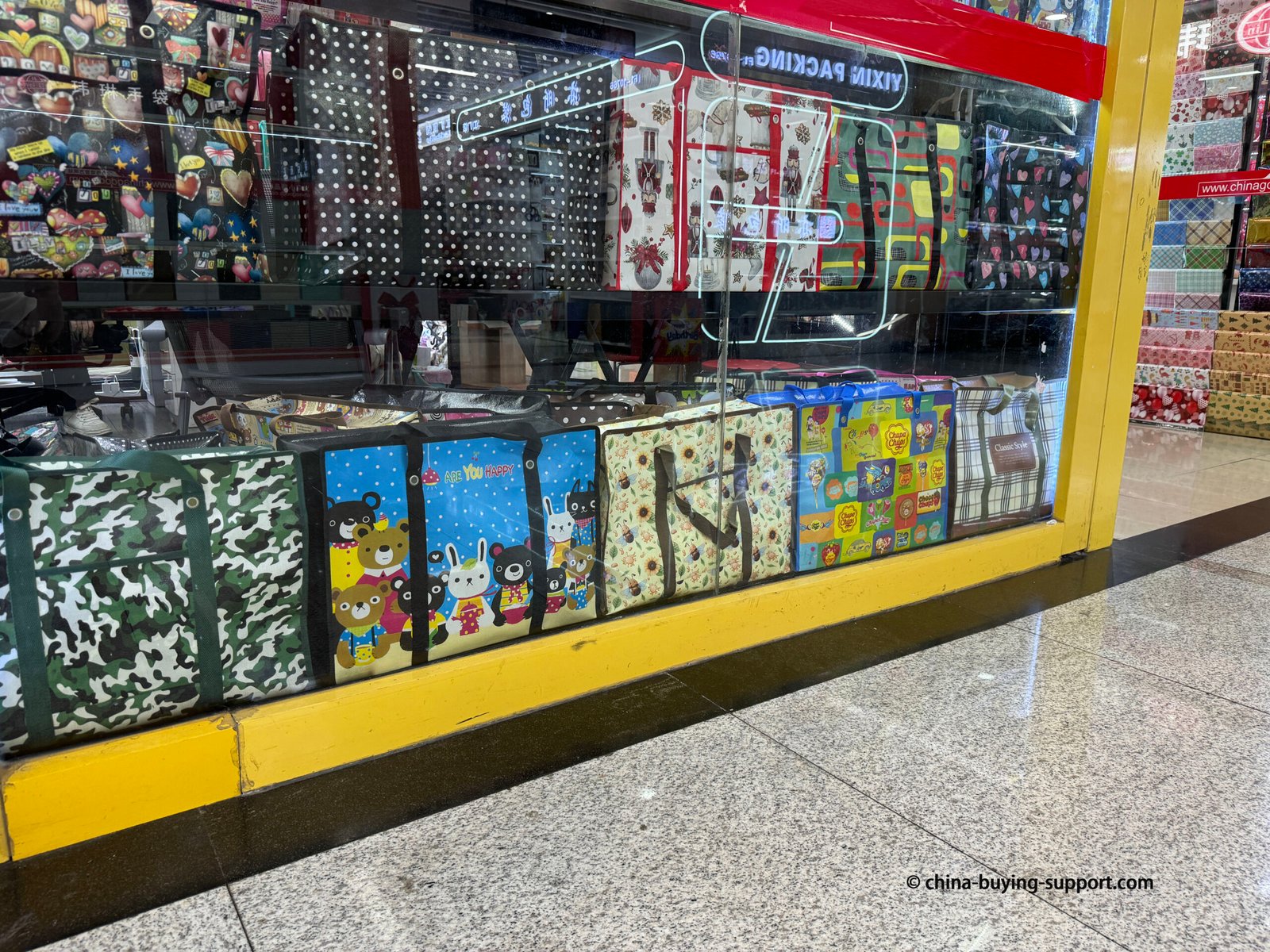 Storefront of a non-woven shopping bag supplier in Yiwu International Trade City District 2, China, displaying large reusable tote bags with colorful prints, cartoon designs, and eco-friendly materials for wholesale buyers.