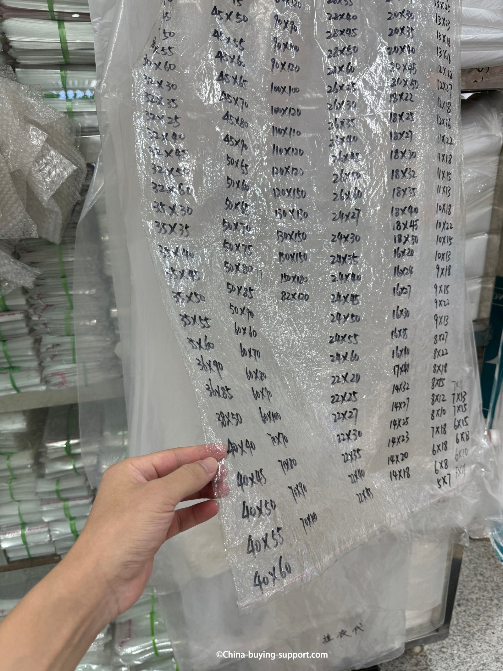 Hand holding a transparent OPP plastic bag size chart showing multiple measurements at a packaging supplier in Yiwu International Trade City Zone 2, suitable for clothing and gift packaging.