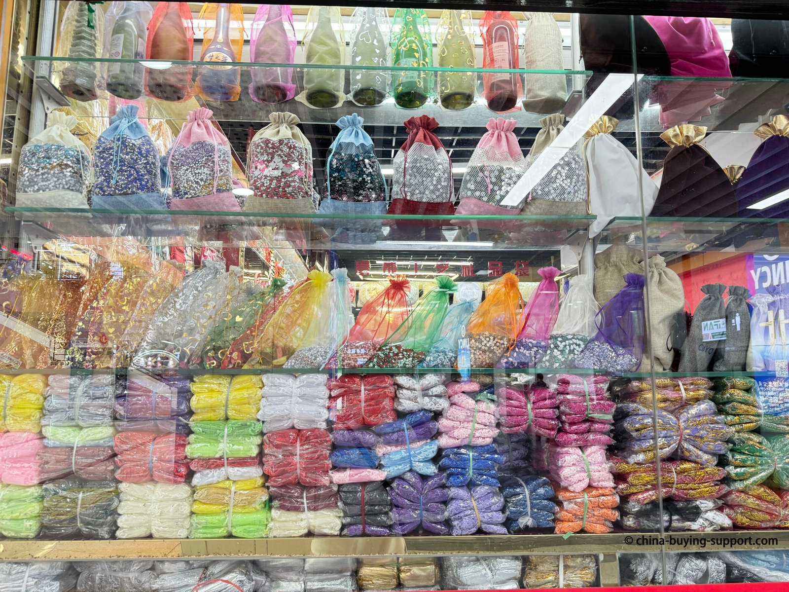 Glass shelf display of colorful organza bags and metallic drawstring pouches at a packaging supplier in Yiwu International Trade City, featuring gift bags used for jewelry, weddings, and party favors.