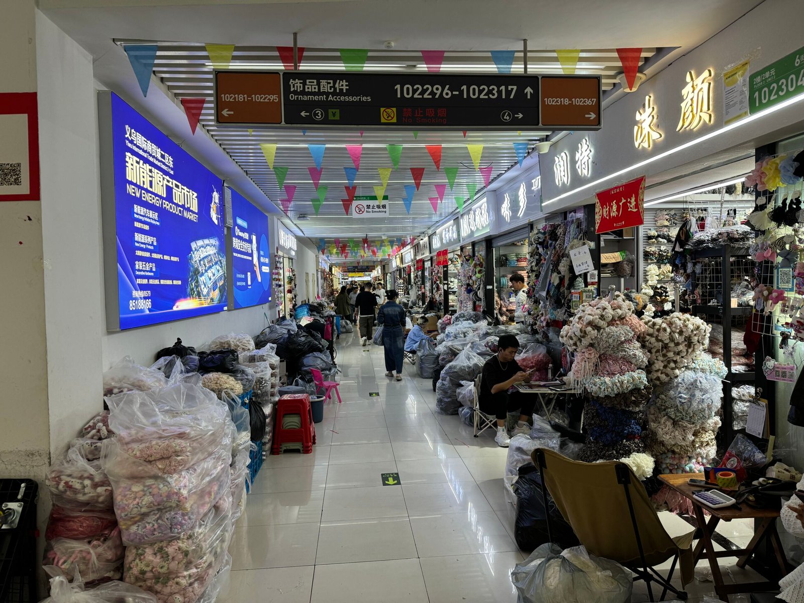 Aisle of the Yiwu ornament accessories market with booth signs 102296–102317, vendors surrounded by bulk bags of hair accessories and fabric decorations.