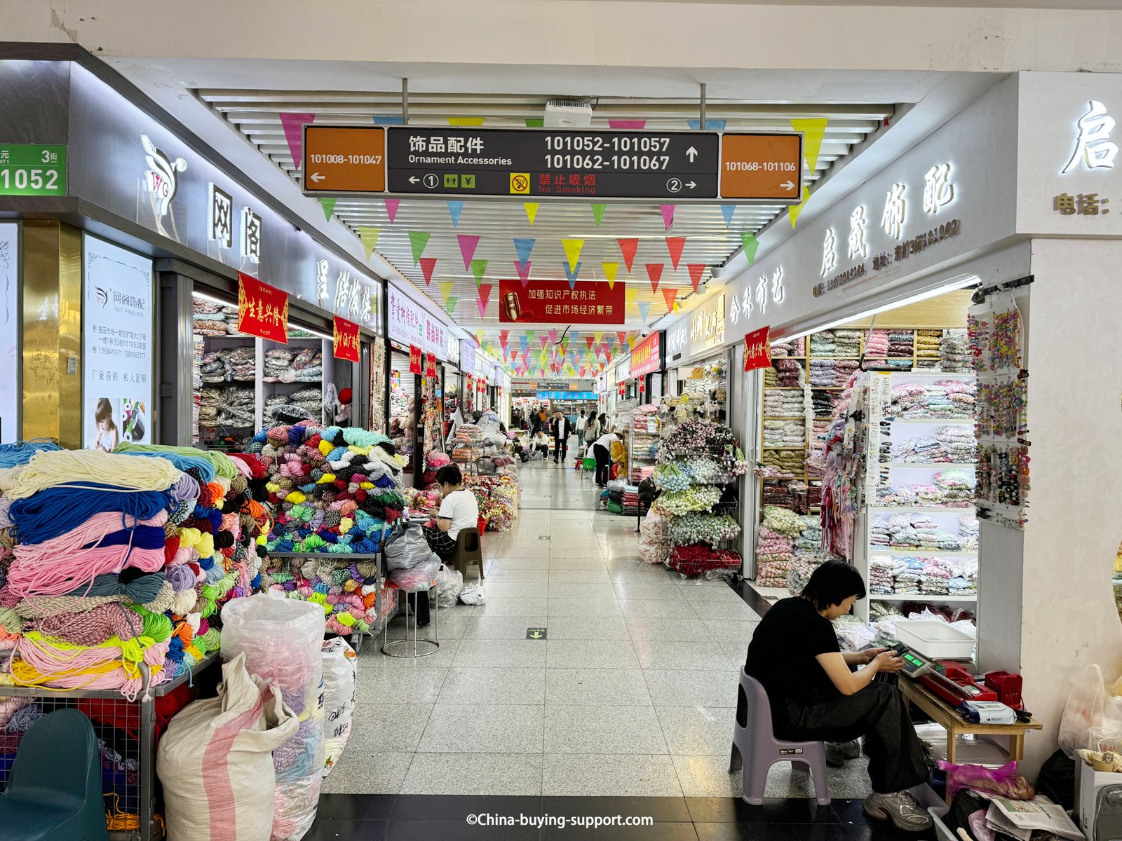 Corridor of the Ornament Accessories area at Yiwu International Trade City with shops selling yarn cords, ribbons, and trims stacked floor-to-ceiling; buyers walking through.