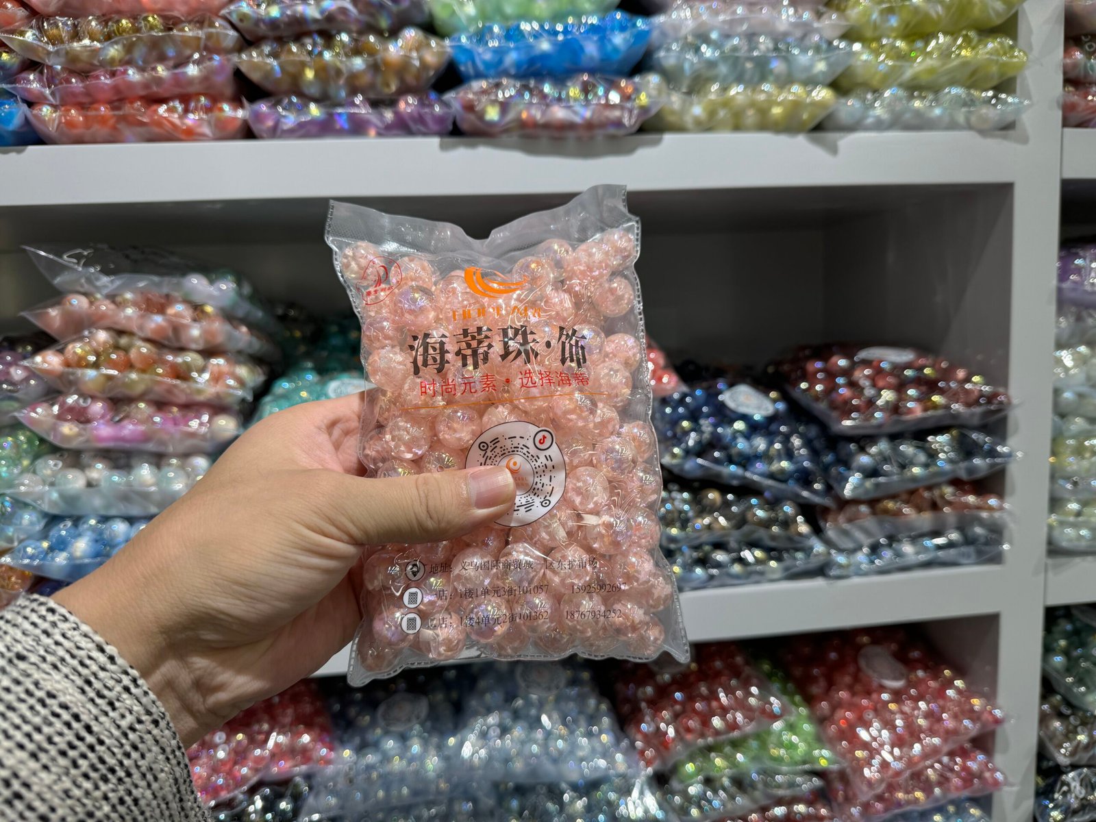Close-up of a hand holding a sealed pack of pink iridescent bubble beads in front of shelves of assorted colors at a Yiwu accessories store.