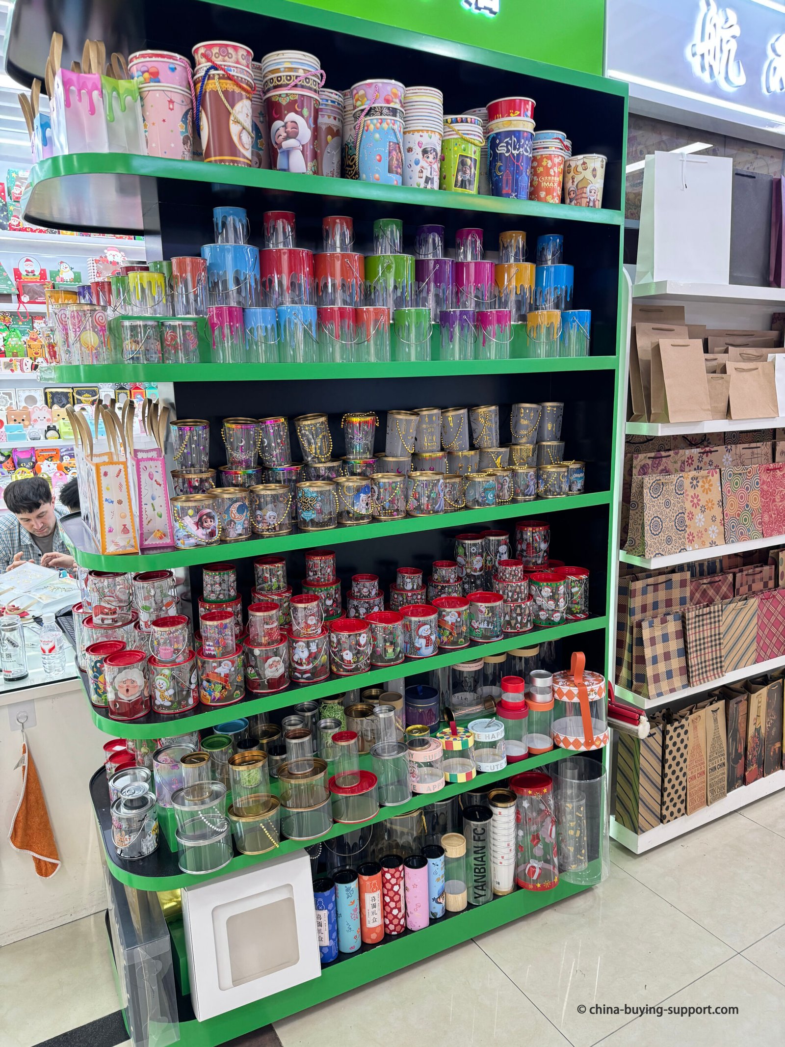 Display shelf filled with colorful plastic gift cans and transparent packaging jars at Yiwu International Trade City District 2 Gift Packaging Market, showing various printed, decorative, and reusable containers for gifts, food, and retail packaging.