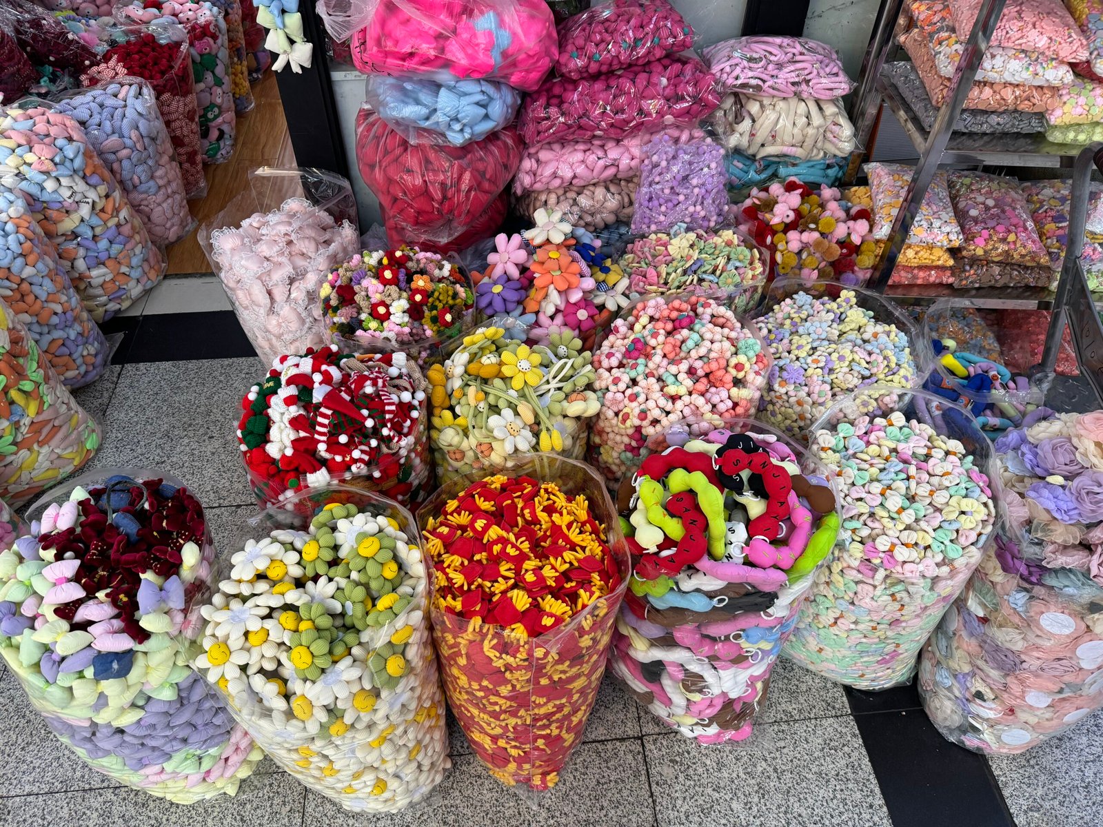 Assortment of plush craft decorations and soft beads in many colors—daisies, hearts, pom-poms—packed in transparent bags at a Yiwu accessories stall.