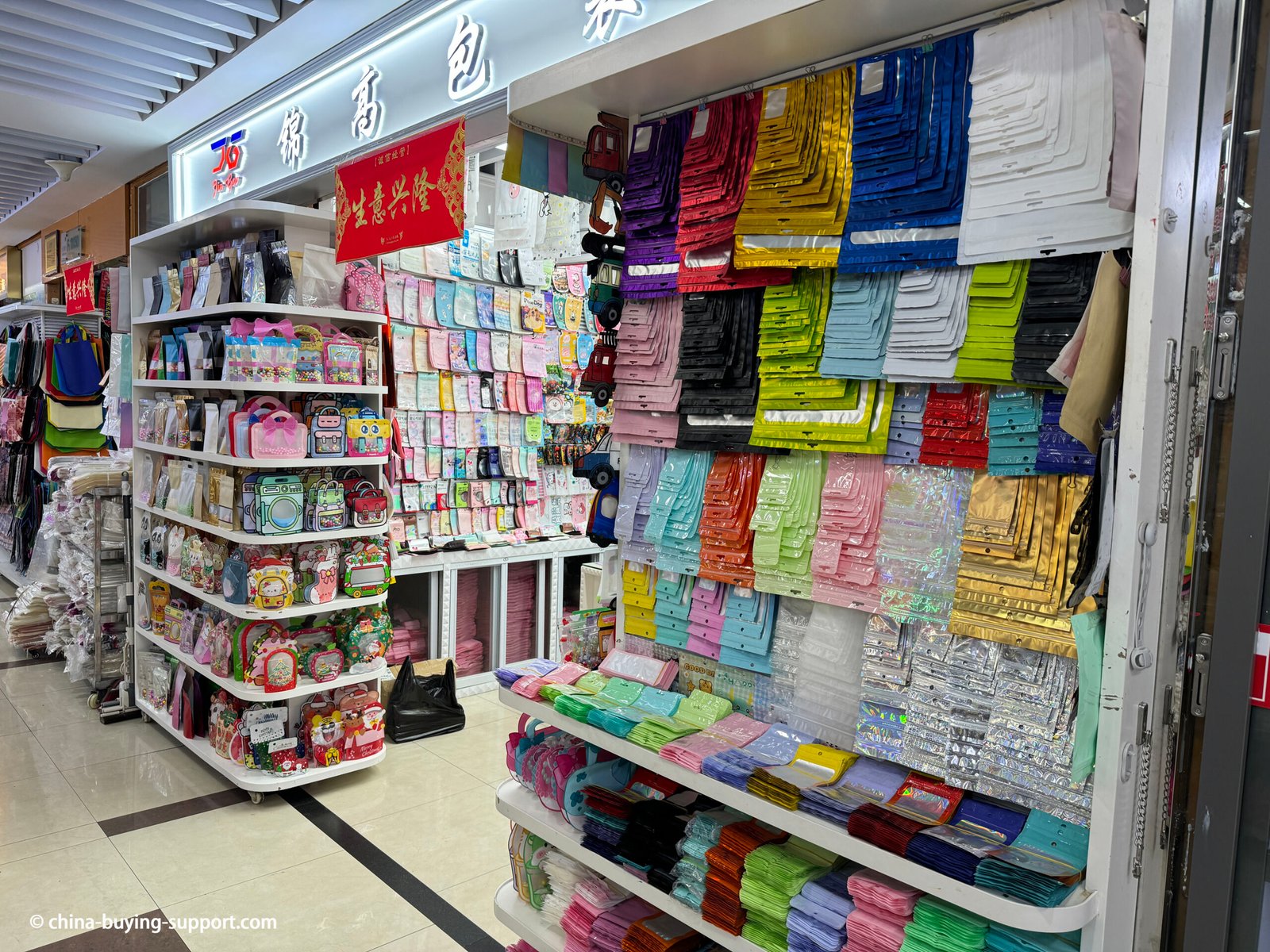 Display shelves at Yiwu International Trade City showing a wide selection of PVC jewelry packaging bags, transparent zip pouches, metallic and holographic gift bags in different colors and sizes for accessories, cosmetics, and retail gift packaging.