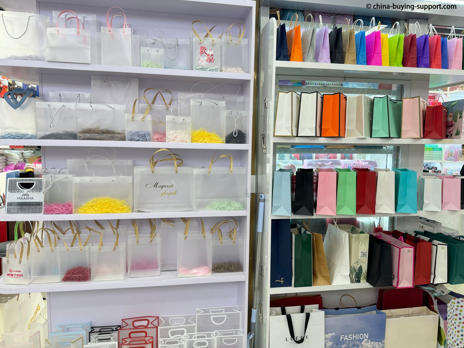 Storefront of a PVC and paper bag supplier in Yiwu International Trade City District 2, China, displaying transparent PVC shopping bags and colorful paper gift bags for wholesale and custom packaging buyers.