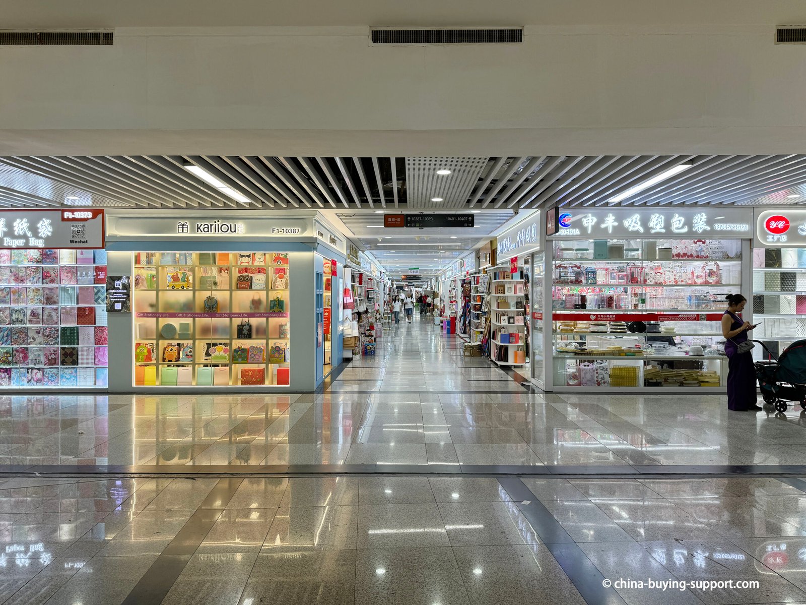 Interior view of Yiwu International Trade City Zone 2 Gift Packaging Market showing the main central aisle with brightly lit shops selling paper bags, PVC packaging, stationery, and gift wrapping materials for retail and wholesale customers.