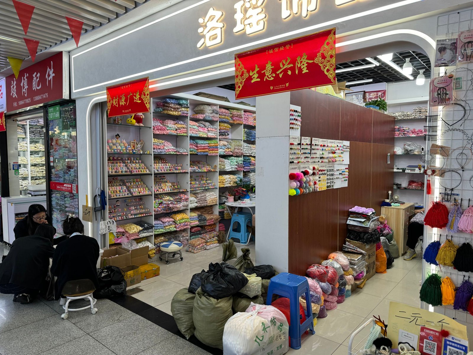 Close-up of Yiwu ornament accessories shop interior with shelves of pastel resin cabochons, flatbacks and bead bags; customers discussing orders on stools.