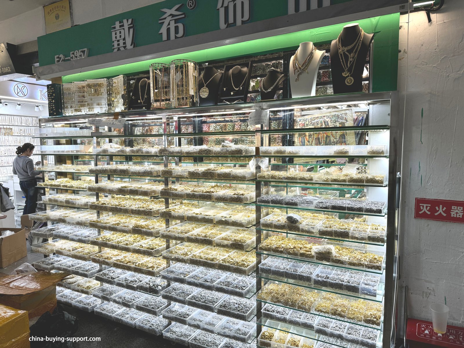 Wide storefront display of gold and silver zinc alloy jewelry findings in Yiwu Market, with trays of pendants, charms, clasps, links and chains arranged on glass shelves.