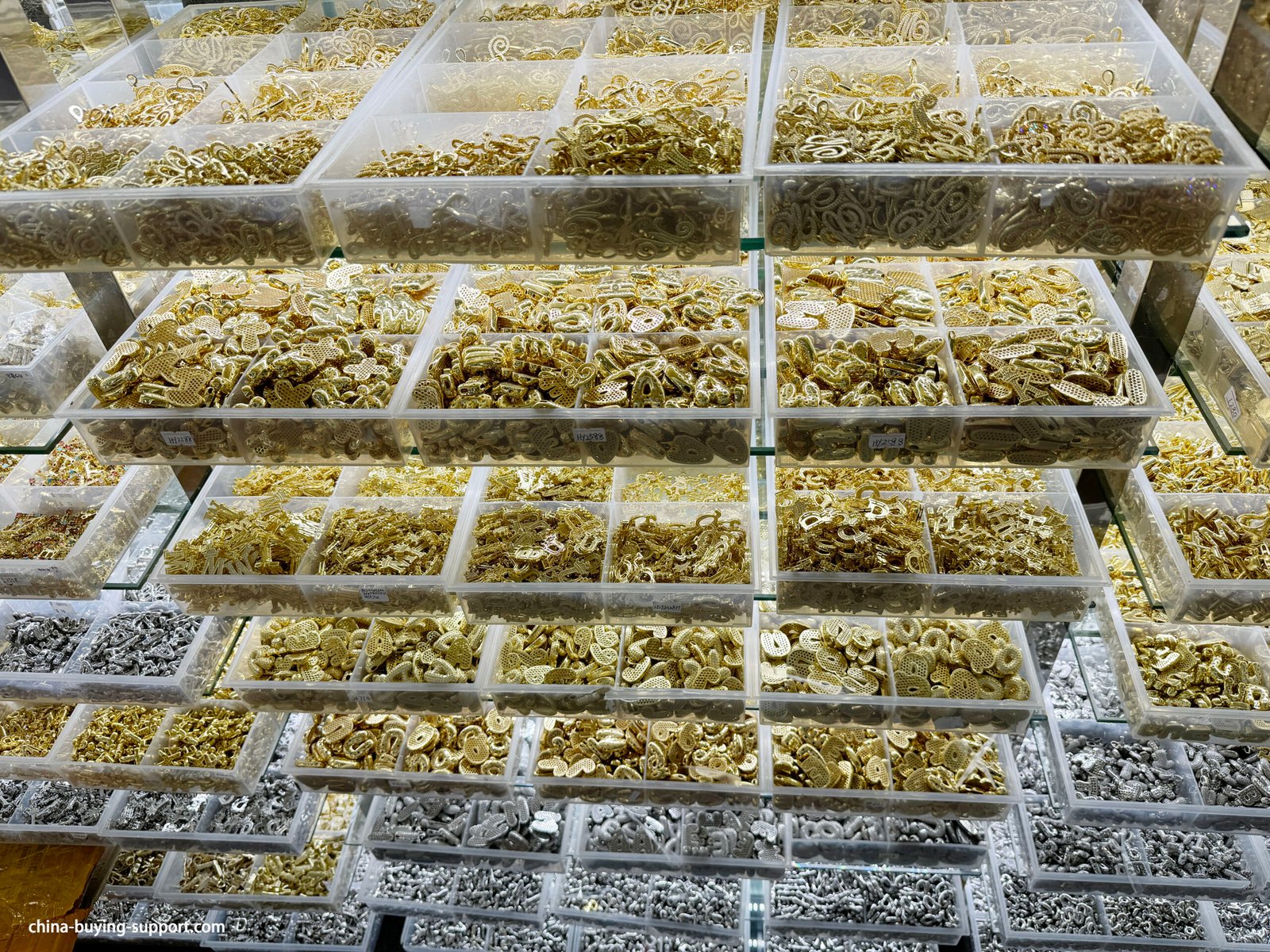 Trays of gold zinc alloy oval links, openwork heart and coin charms, mixed connectors, and silver findings on lower shelves in Yiwu accessories market.