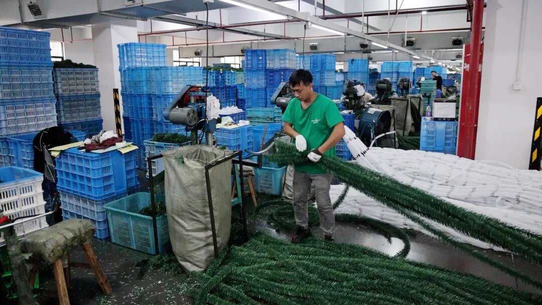 Factory worker in a Yiwu Christmas tree plant assembling long green artificial tree branches beside machines and tall stacks of blue plastic crates.
