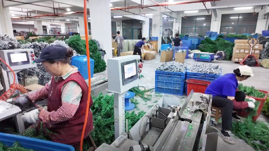 Workers in a Yiwu artificial Christmas tree factory assembling green PVC branches at machines and sorting parts into blue plastic crates and cartons on the workshop floor.