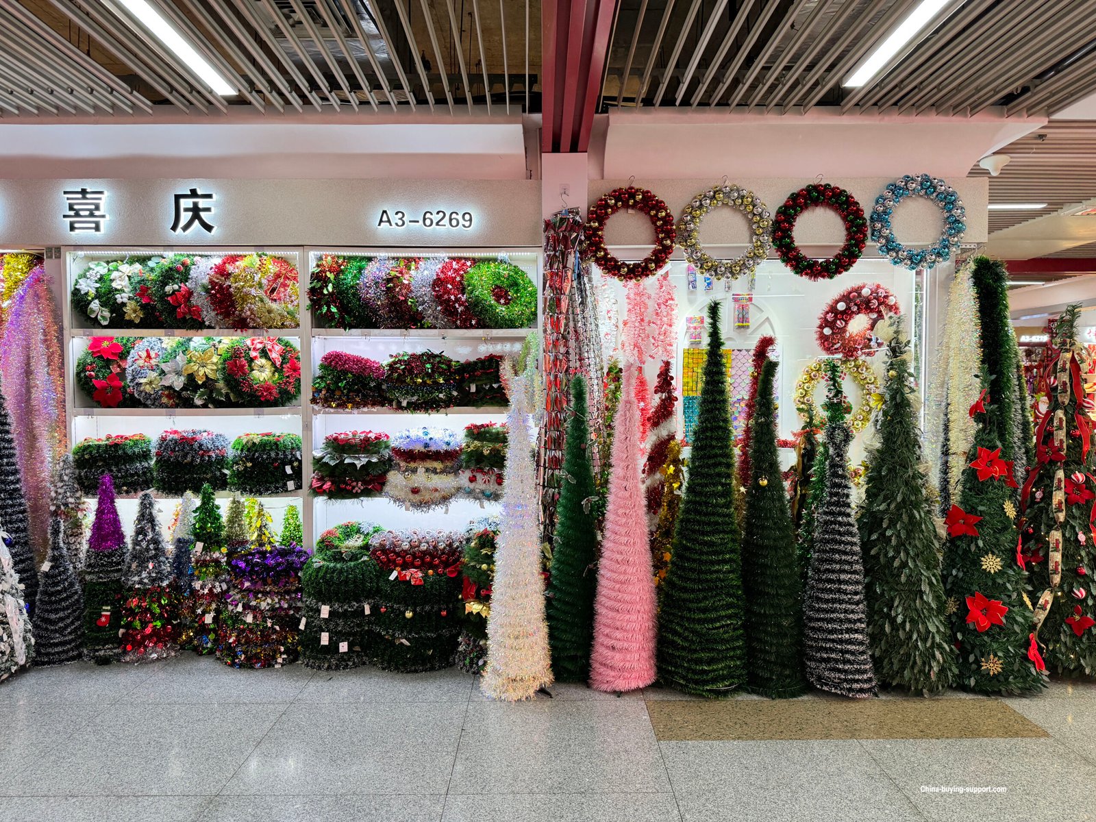Yiwu Christmas wholesale market stall A3-6269 with rows of cone-shaped artificial Christmas trees, tinsel garlands and bright wreaths on white shelves.