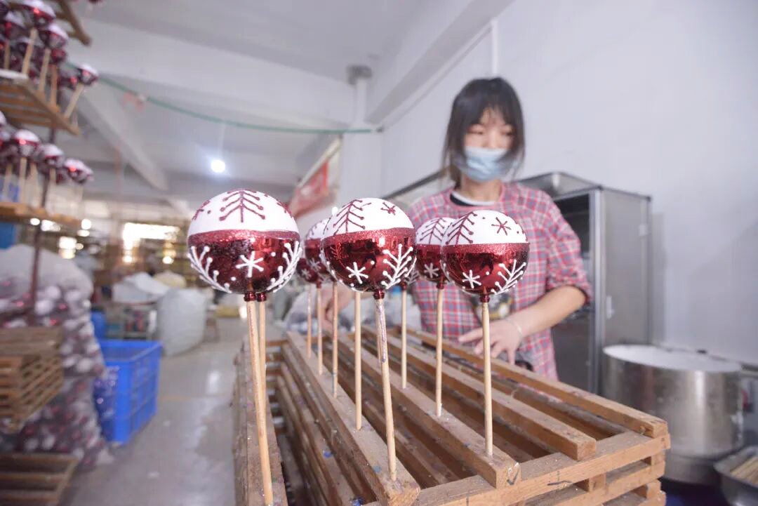 Chinese factory worker wearing a mask carefully making red and white glitter Christmas baubles with snowflake patterns on wooden sticks inside a Yiwu Christmas ornament workshop.