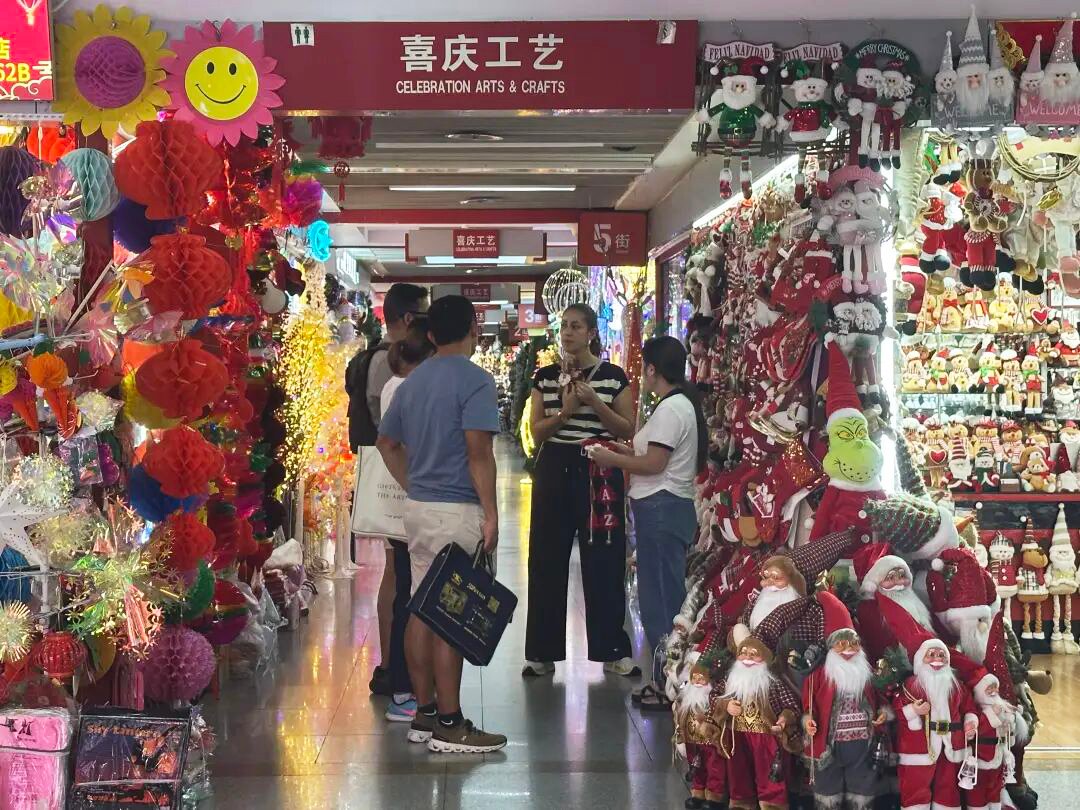 Group of foreign buyers talking with Yiwu Christmas decoration suppliers in a colorful market aisle filled with Santa dolls, festive crafts and bright lights.
