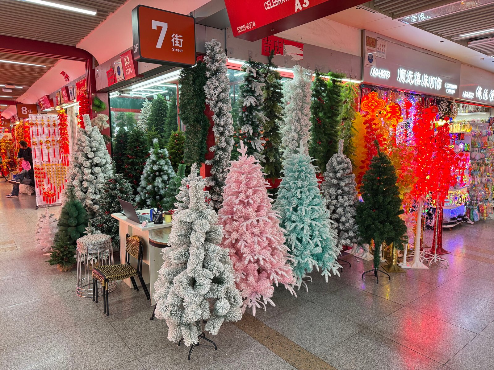 A variety of flocked artificial Christmas trees standing in front of a stall at the Yiwu wholesale market, including white, pastel pink, icy blue, mint green and dark green trees, with a small desk, stools and mirrors reflecting more trees inside.