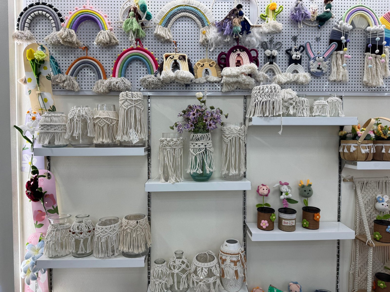 Macrame-covered glass jars and colorful crochet rainbow wall hangings on white shelves in a Yiwu home decor market shop.