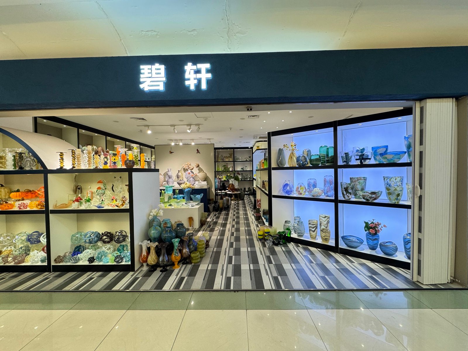 Wide storefront of a Chinese art glass home decor shop at Yiwu market showing illuminated shelves on both sides filled with colorful vases and bowls leading into a deep showroom.