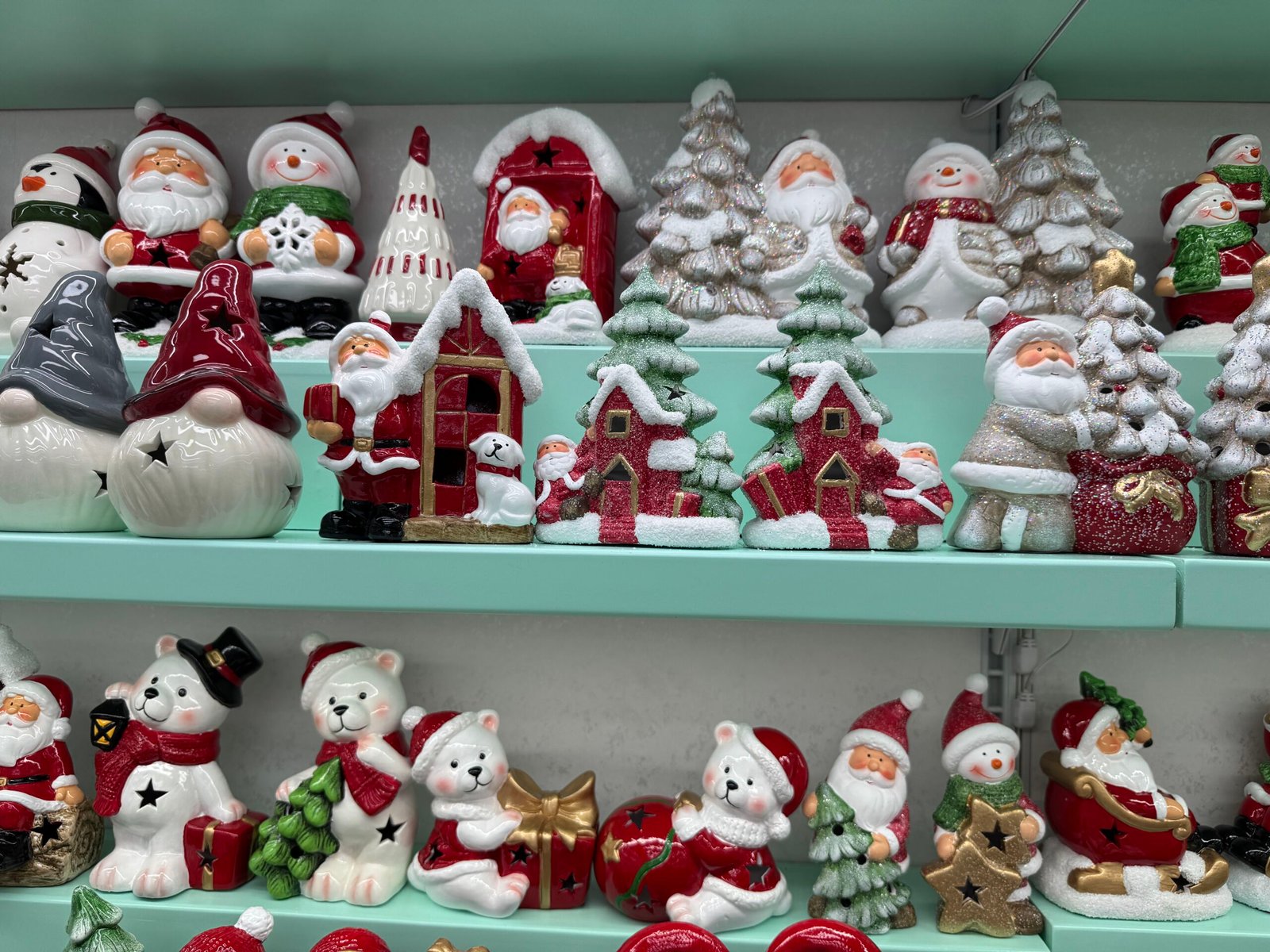 Rows of glossy ceramic Christmas ornaments on mint-green shelves in the Yiwu wholesale market, including Santa Claus figures, smiling snowmen, glittery Christmas trees, red snow village houses and polar bear decorations covered with artificial snow.