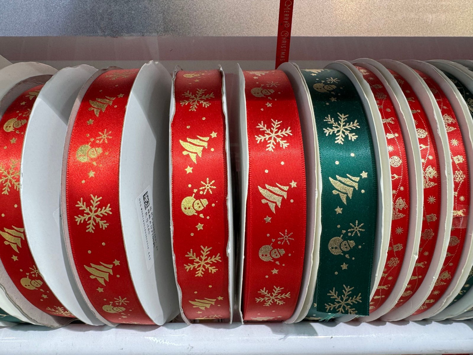 Several rolls of red and green satin Christmas ribbon in Yiwu, China, each printed with gold snowflakes, Christmas trees, stars and snowman icons, neatly lined up on white cardboard spools.