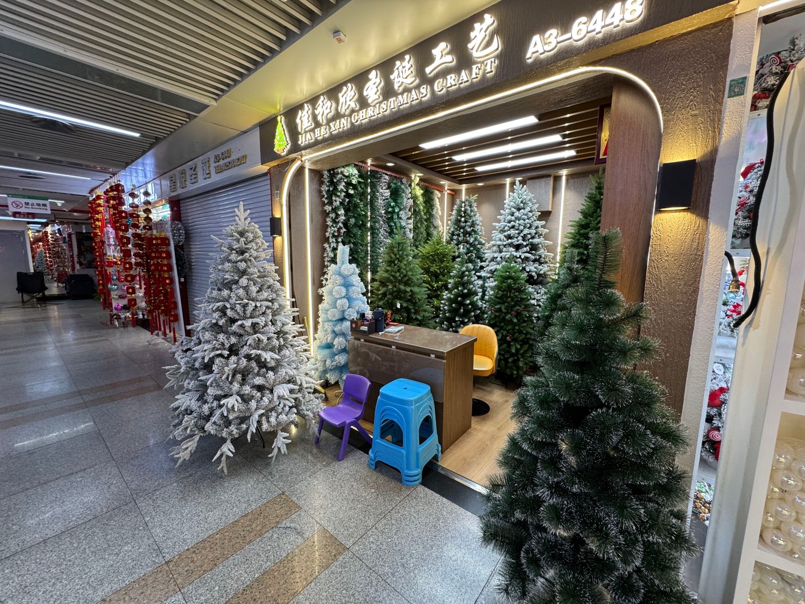 Storefront of the Jia He Xin Christmas Craft shop in the Yiwu Christmas decoration wholesale market, with tall flocked white, blue and green artificial Christmas trees arranged around a small counter and plastic stools at the entrance.