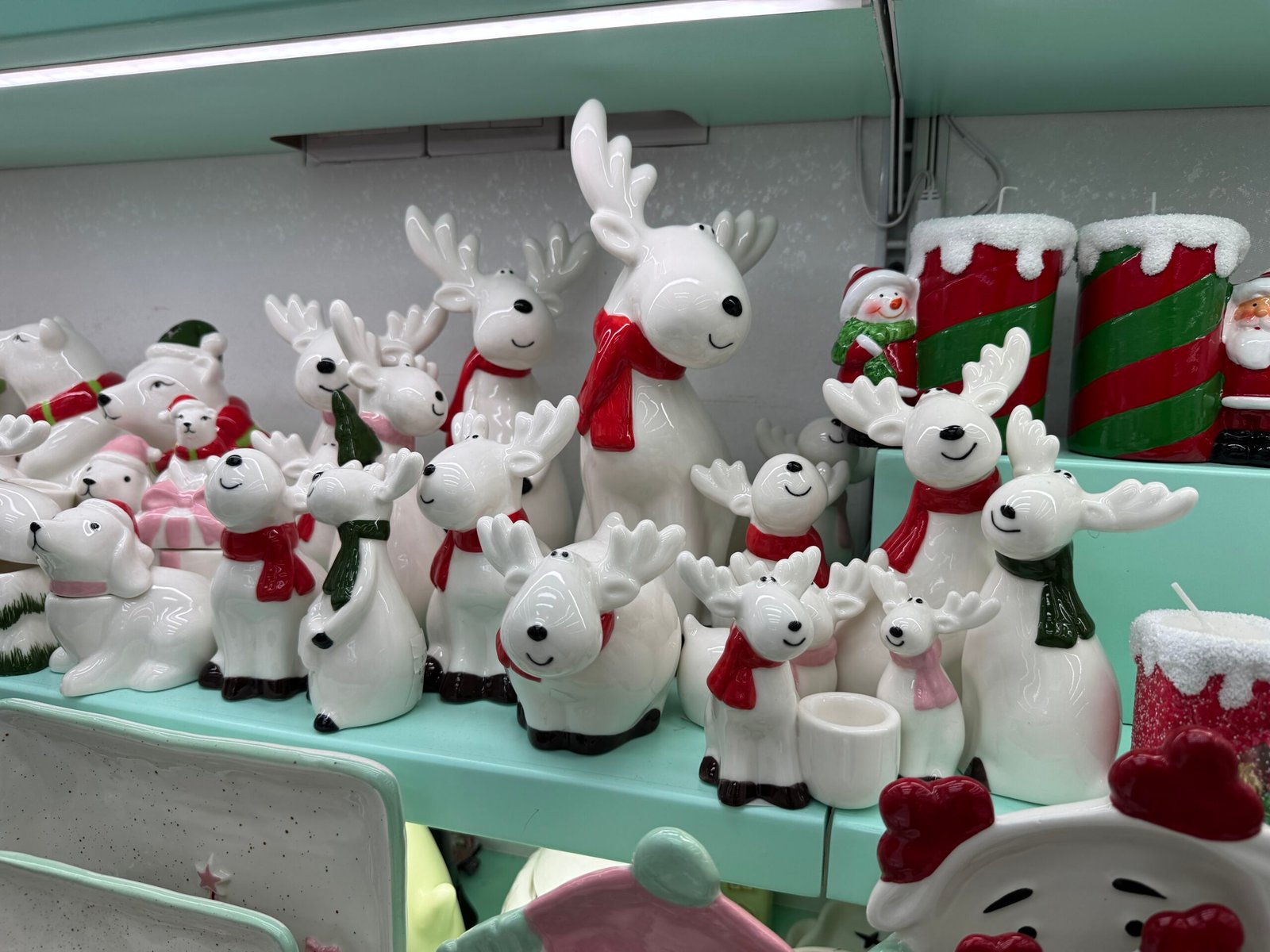 Display of many white ceramic reindeer and polar bear figurines in different sizes at the Yiwu Christmas wholesale market, all with smiling faces and red or green scarves, surrounded by other festive decor such as striped red-and-green candle holders and snowman ornaments.