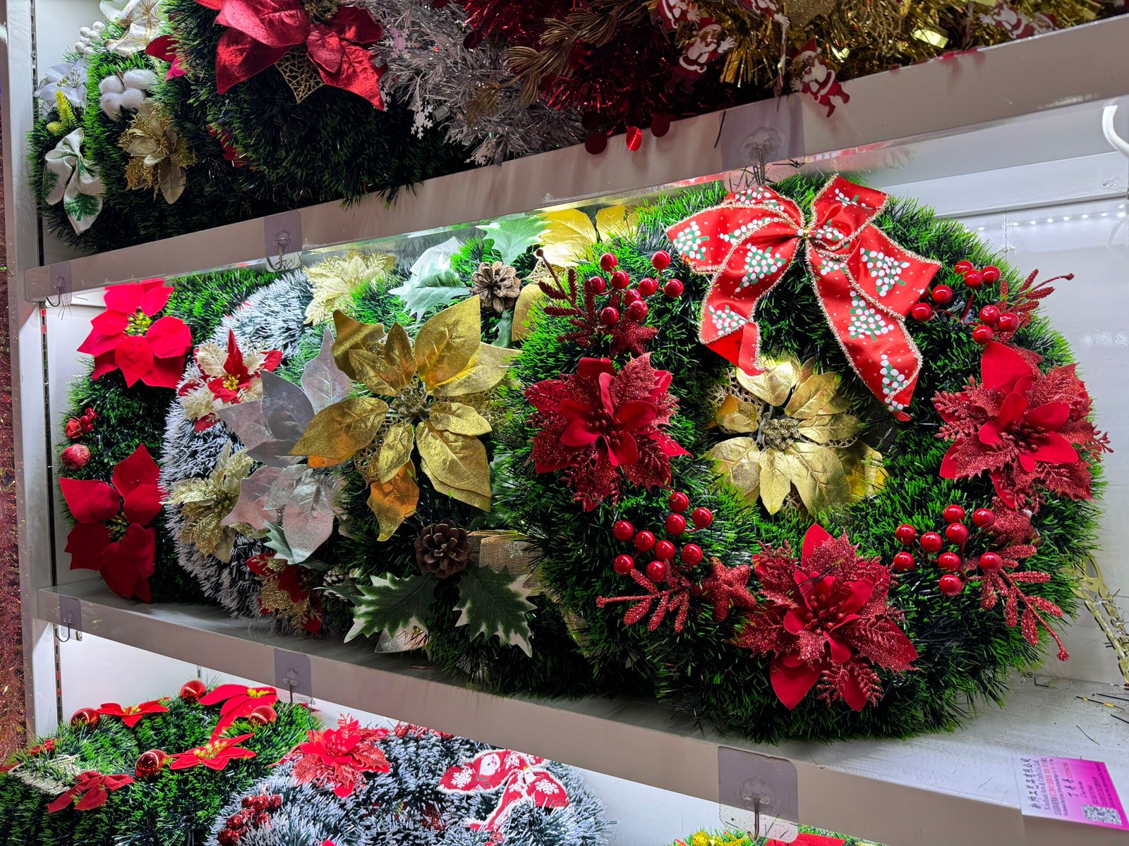 Green artificial Christmas wreaths on a shelf in the Yiwu wholesale market, richly decorated with red poinsettia flowers, gold and silver leaves, pinecones, red berries and a large red ribbon bow.