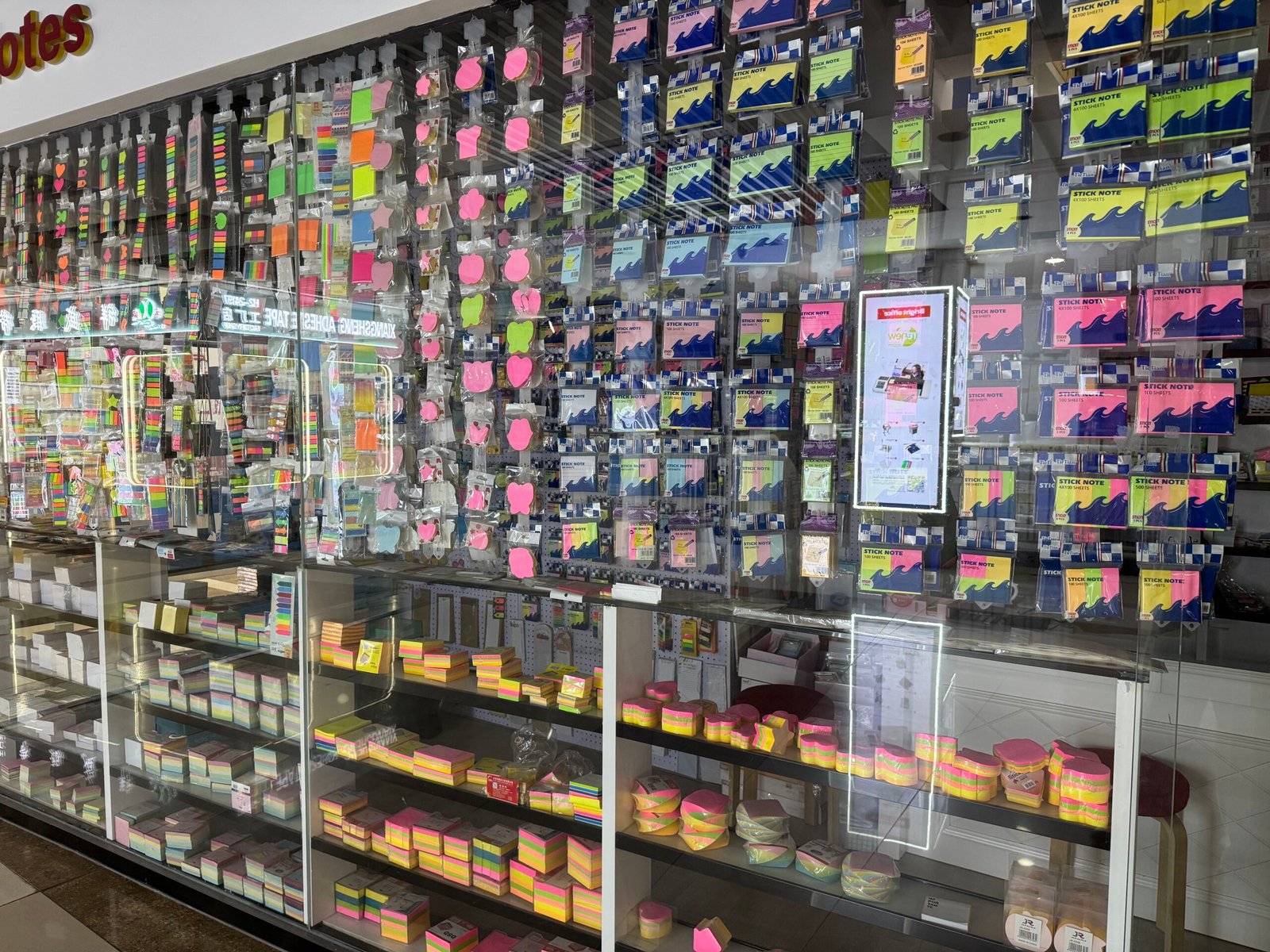 Wall of assorted sticky notes, index tabs and shaped memo pads in bright colors with additional packs stored in glass cabinets at a Yiwu stationery shop.