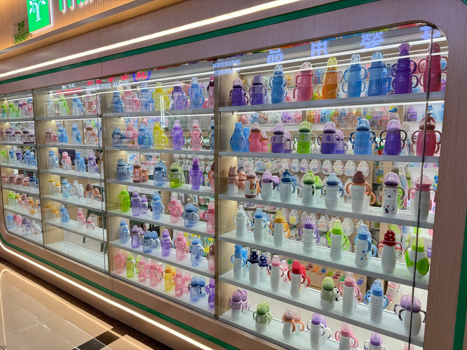 Large glass display wall in Yiwu International Trade City District 4 showing multiple shelves of baby bottles and sippy cups in pastel colors with handles and measurement markings.