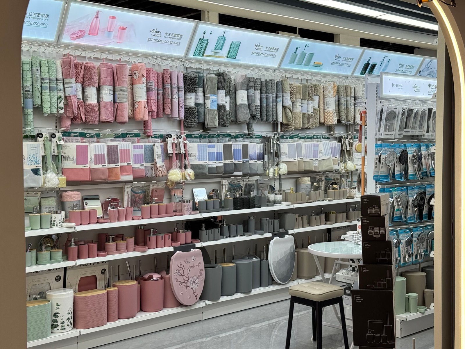 Interior wall of a bathroom accessories shop in Yiwu District 4 displaying rolled bath mats, shower curtains, storage bags and matching bathroom sets in pink, grey and neutral colors.