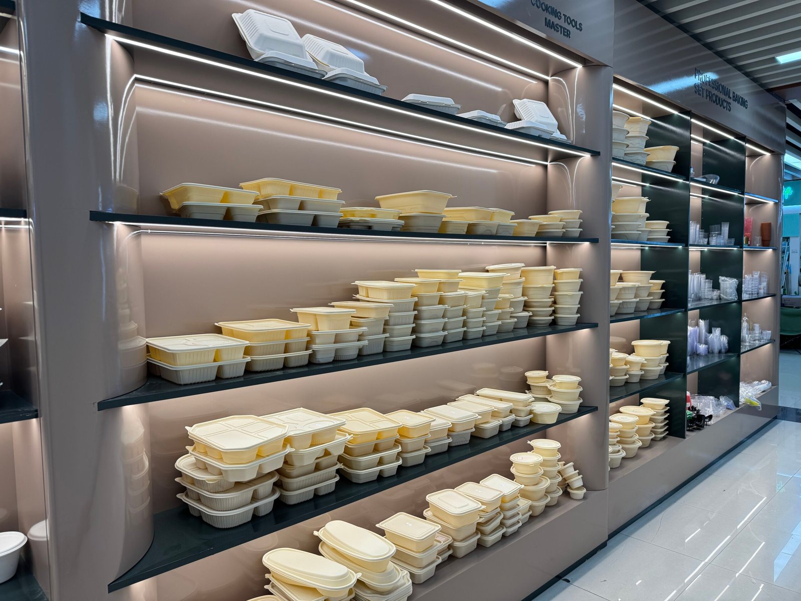 Sample wall of a biodegradable disposable tableware factory in Yiwu International Trade City District 4, with rows of cream-colored compostable lunch boxes and food containers stacked on modern backlit shelves.