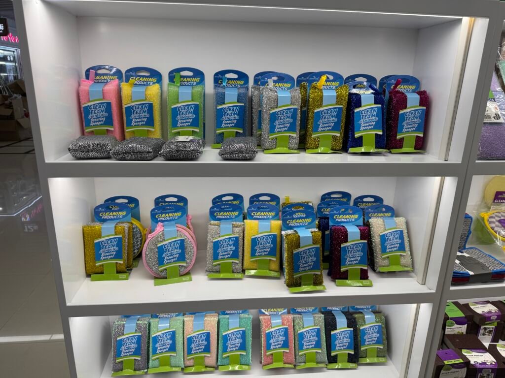 Rows of glitter metallic scouring sponges and cleaning pads in pink, green, silver and gold packaging displayed on white shelves in a Yiwu International Trade City District 4 shop.