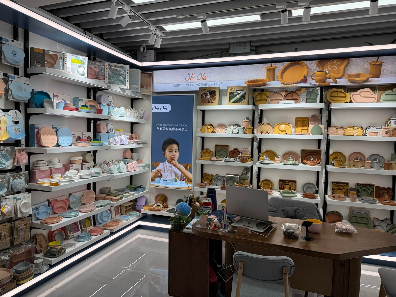Ole Ola kids silicone tableware shop in Yiwu District 4 with shelves full of pastel plates, bowls and cups surrounding a poster of a child eating.