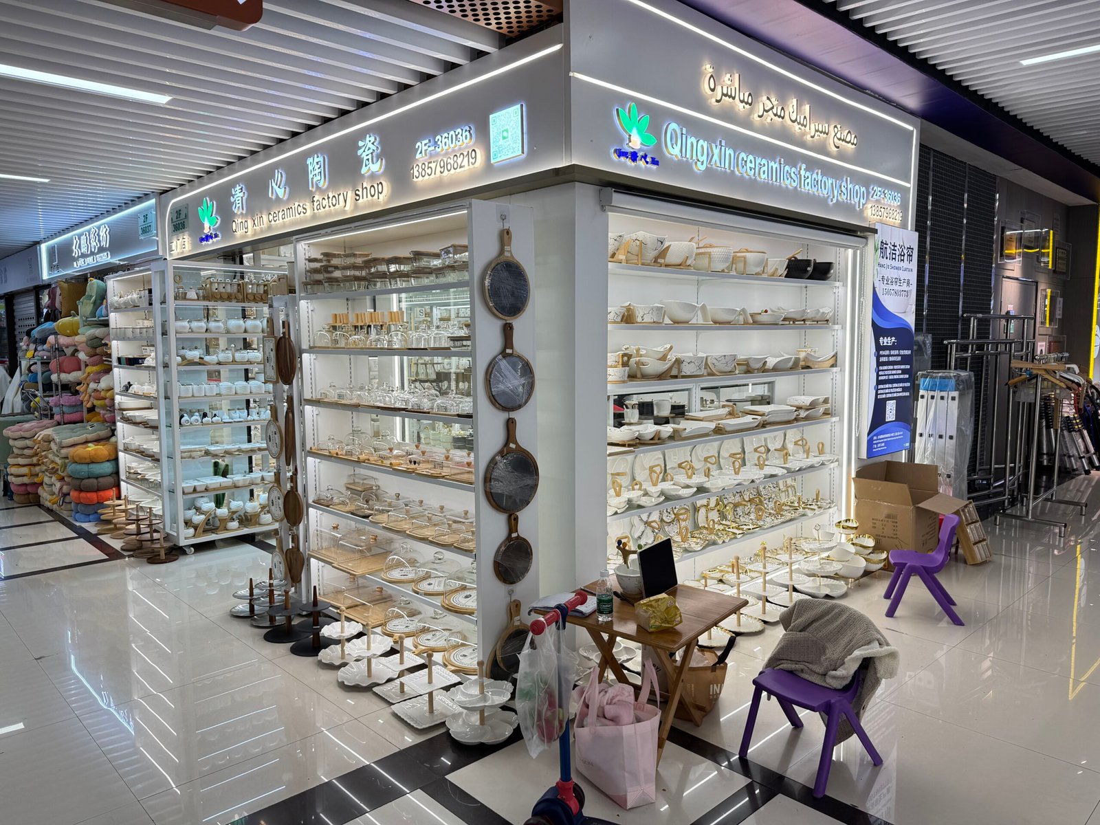 Corner view of Qingxin ceramics factory shop in Yiwu International Trade City District 4 with bright shelves of ceramic plates, bowls, trays and glass bottles on bamboo stands.