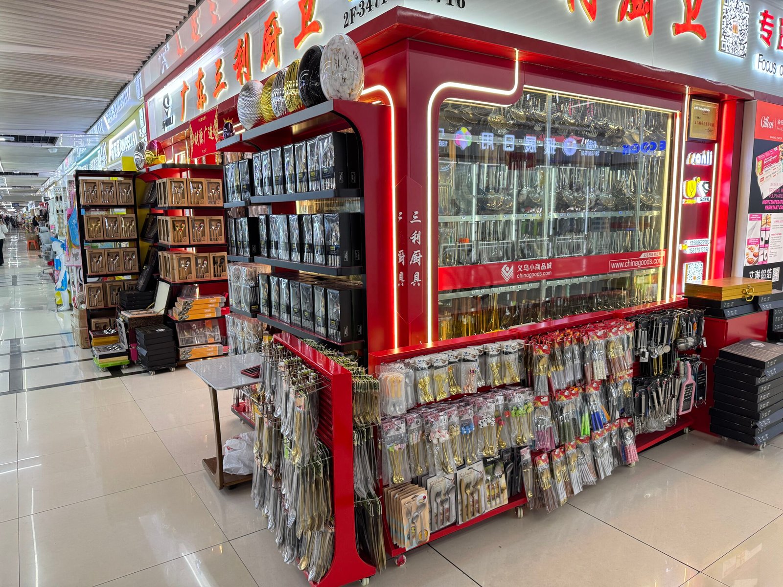Red-themed kitchen cutlery factory shop in Yiwu District 4 with hanging stainless steel spoons and forks in glass cabinets and blister-carded utensil sets on front racks.
