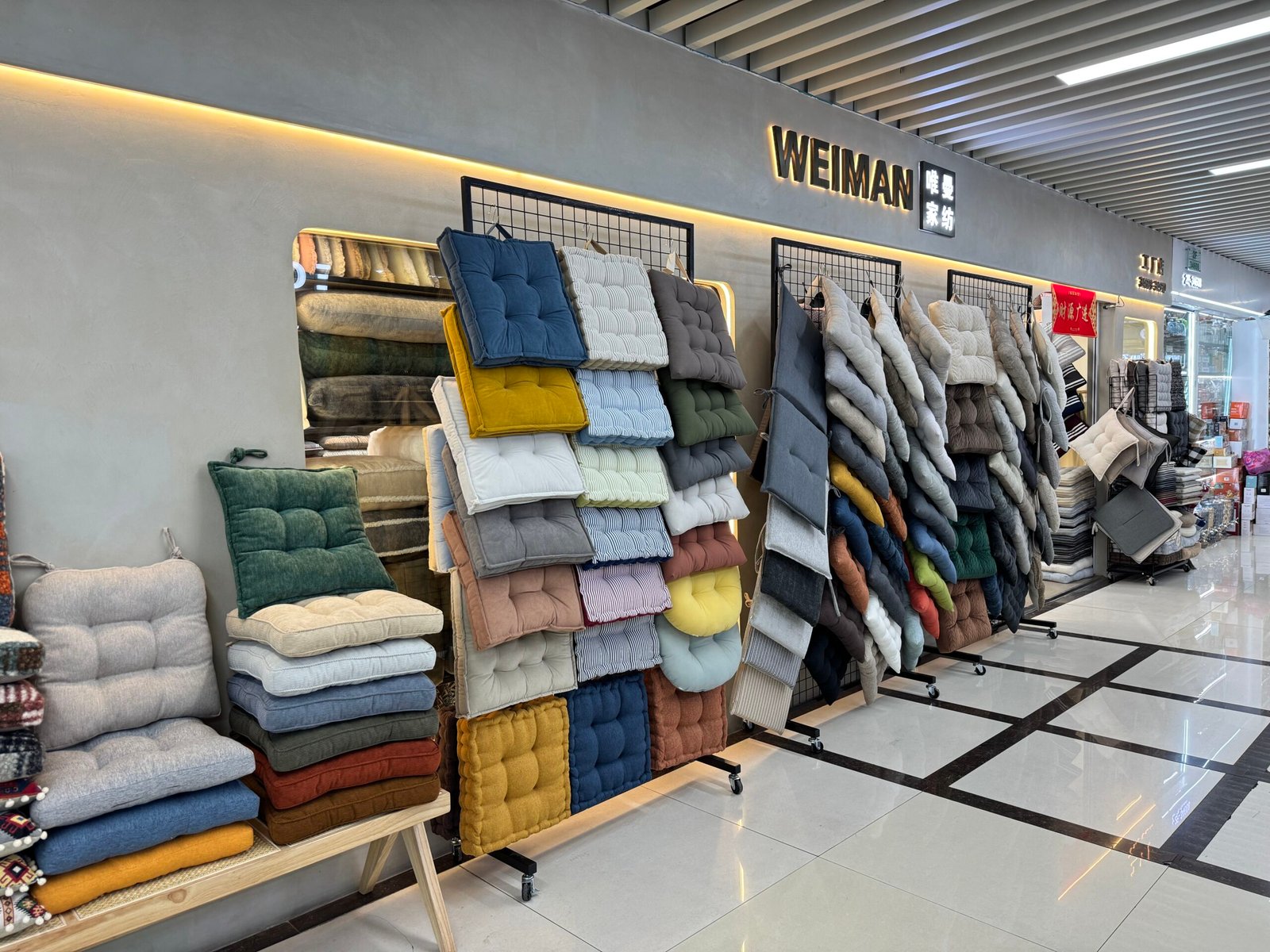 Rows of thick seat cushions and chair pads in various colors and fabrics hanging on racks in front of the WEIMAN shop at Yiwu International Trade City District 4 daily necessities area.