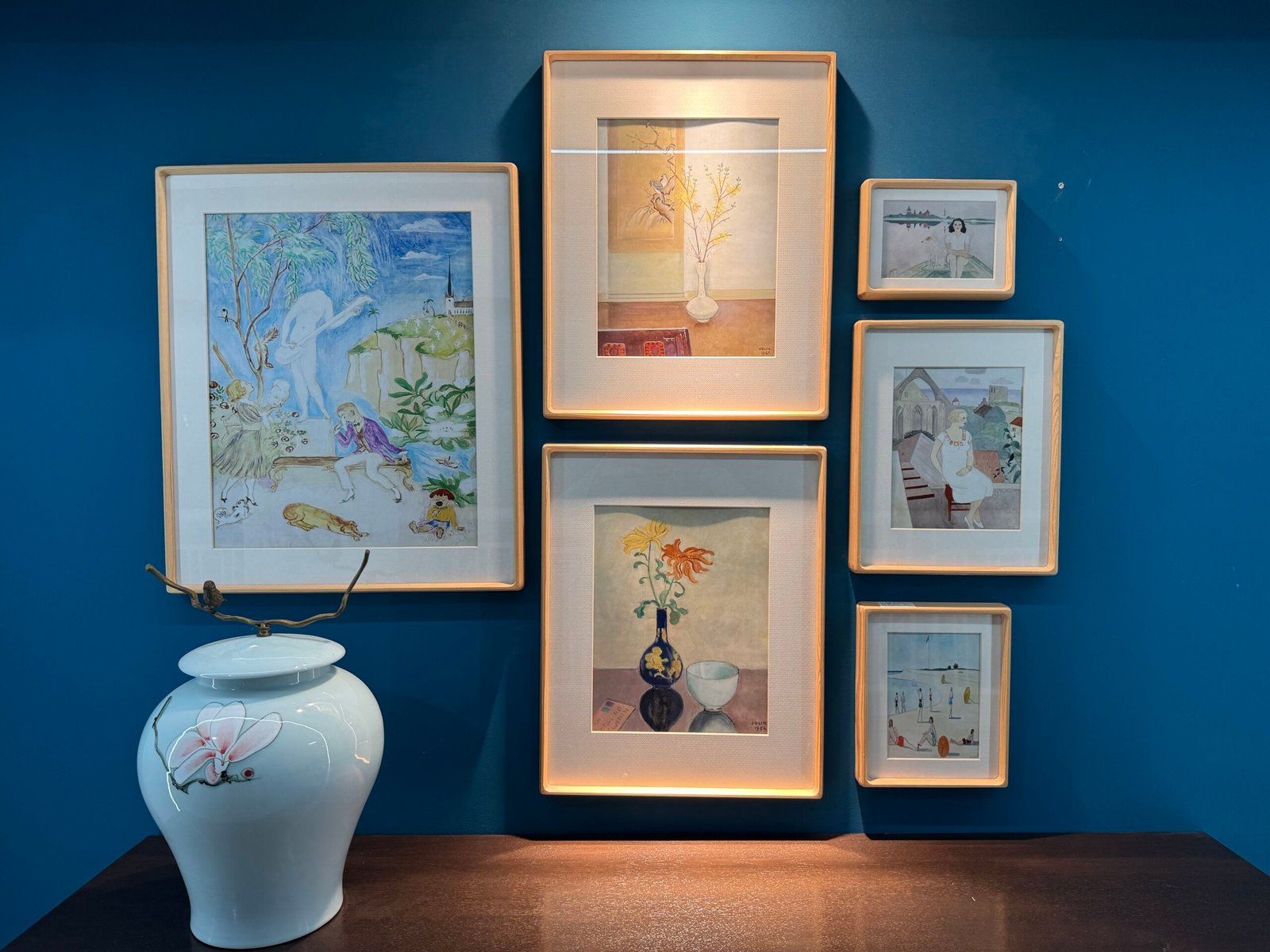Arrangement of six framed art prints in different sizes with warm lighting, mounted on a teal wall above a white ceramic vase in a Yiwu International Trade City frame store.