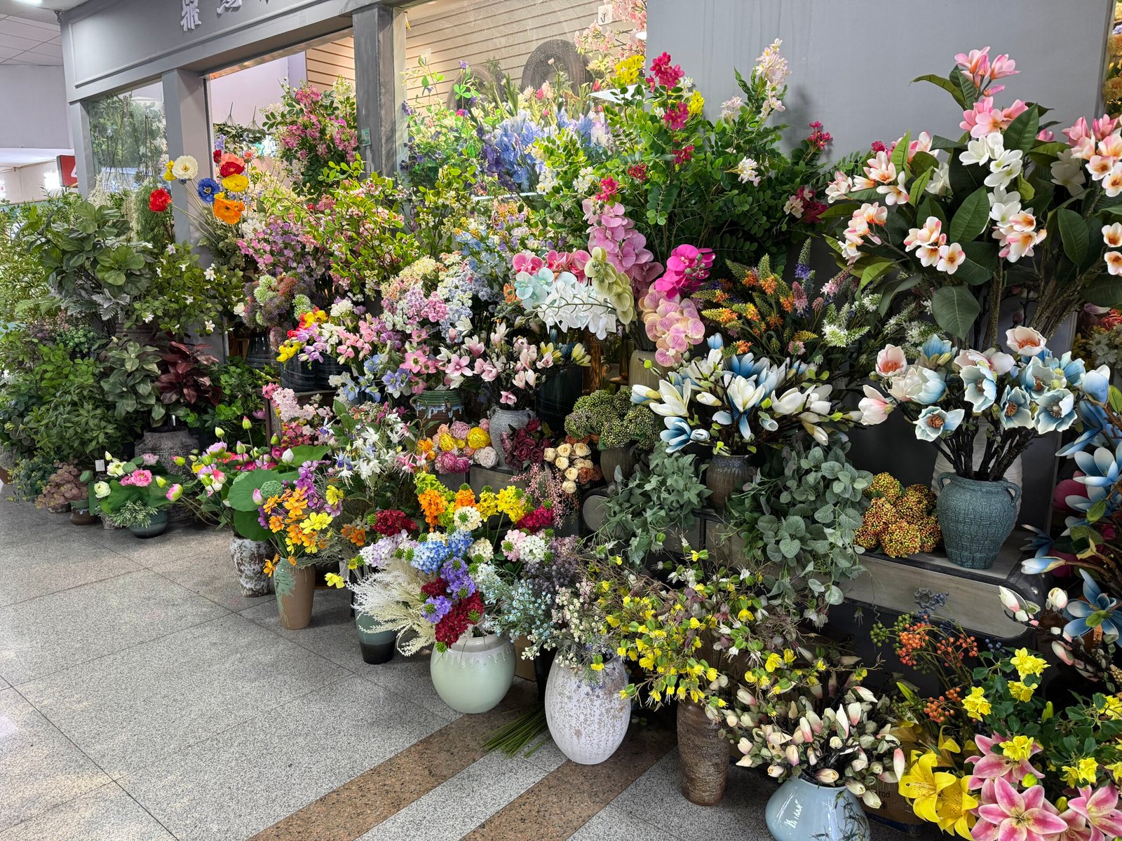 Front of an artificial flower shop in Yiwu home decoration market packed with faux orchids, lilies, bushes and greenery in decorative pots and planters.