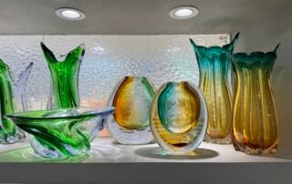Assorted green, amber, and teal art glass vases and bowls on a lit shelf in the Yiwu home decor market in China.