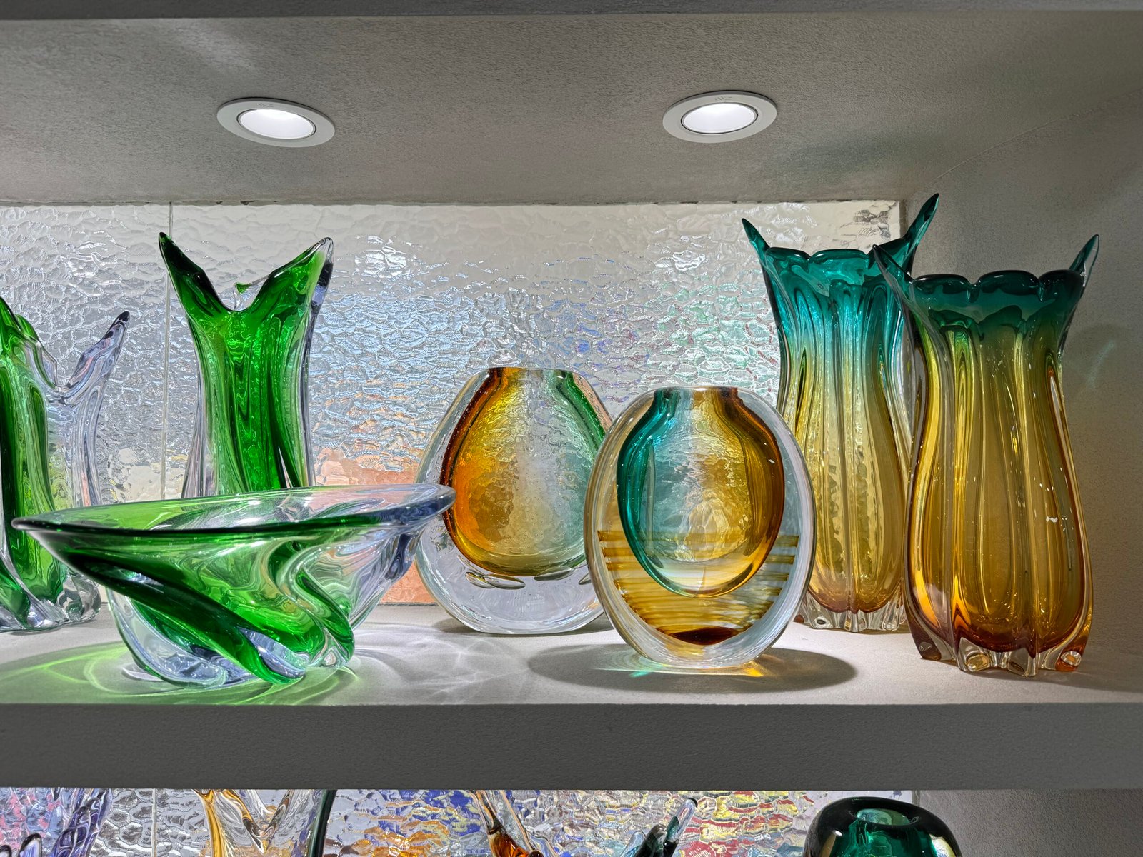Assorted green, amber, and teal art glass vases and bowls on a lit shelf in the Yiwu home decor market in China.