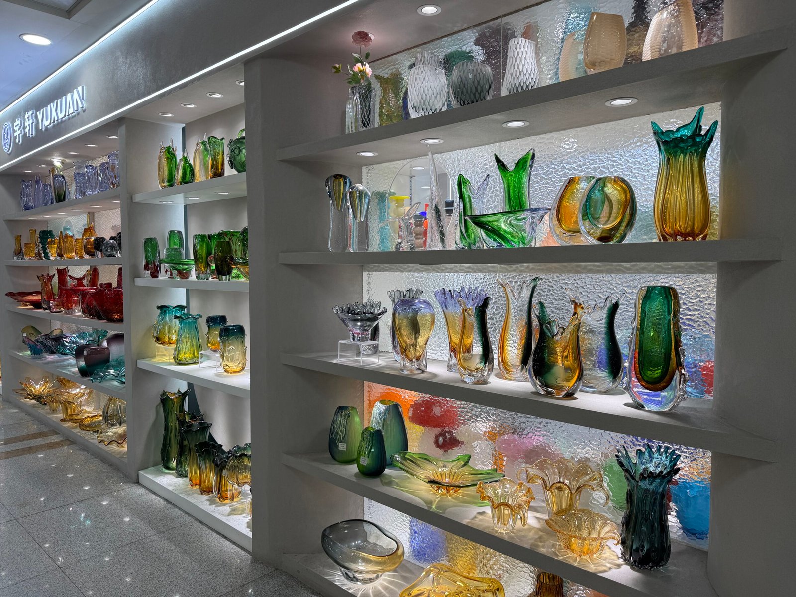 Display of handcrafted style art glass vases and bowls in green, yellow and blue on white shelves against a textured back wall at Yiwu home decoration market.