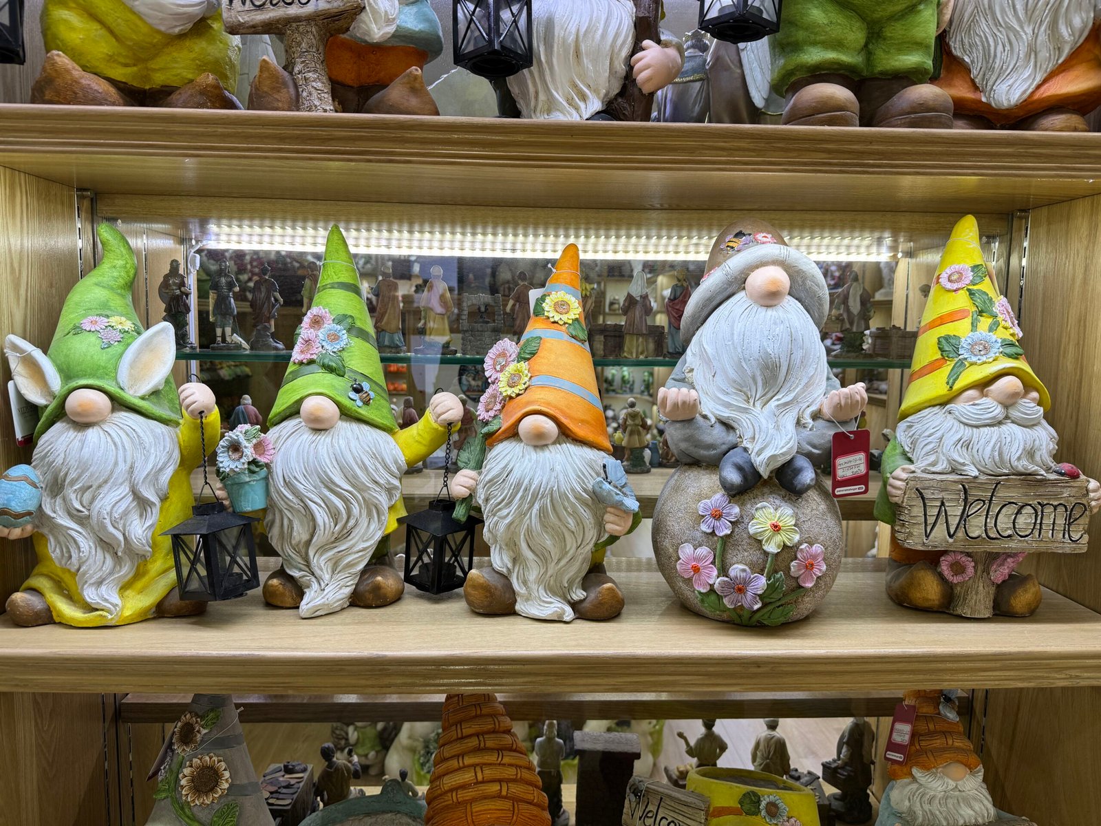 Five colorful resin garden gnome statues with tall flower hats, white beards, lanterns and a wooden “Welcome” sign displayed on a shelf at a Yiwu home decoration market store.