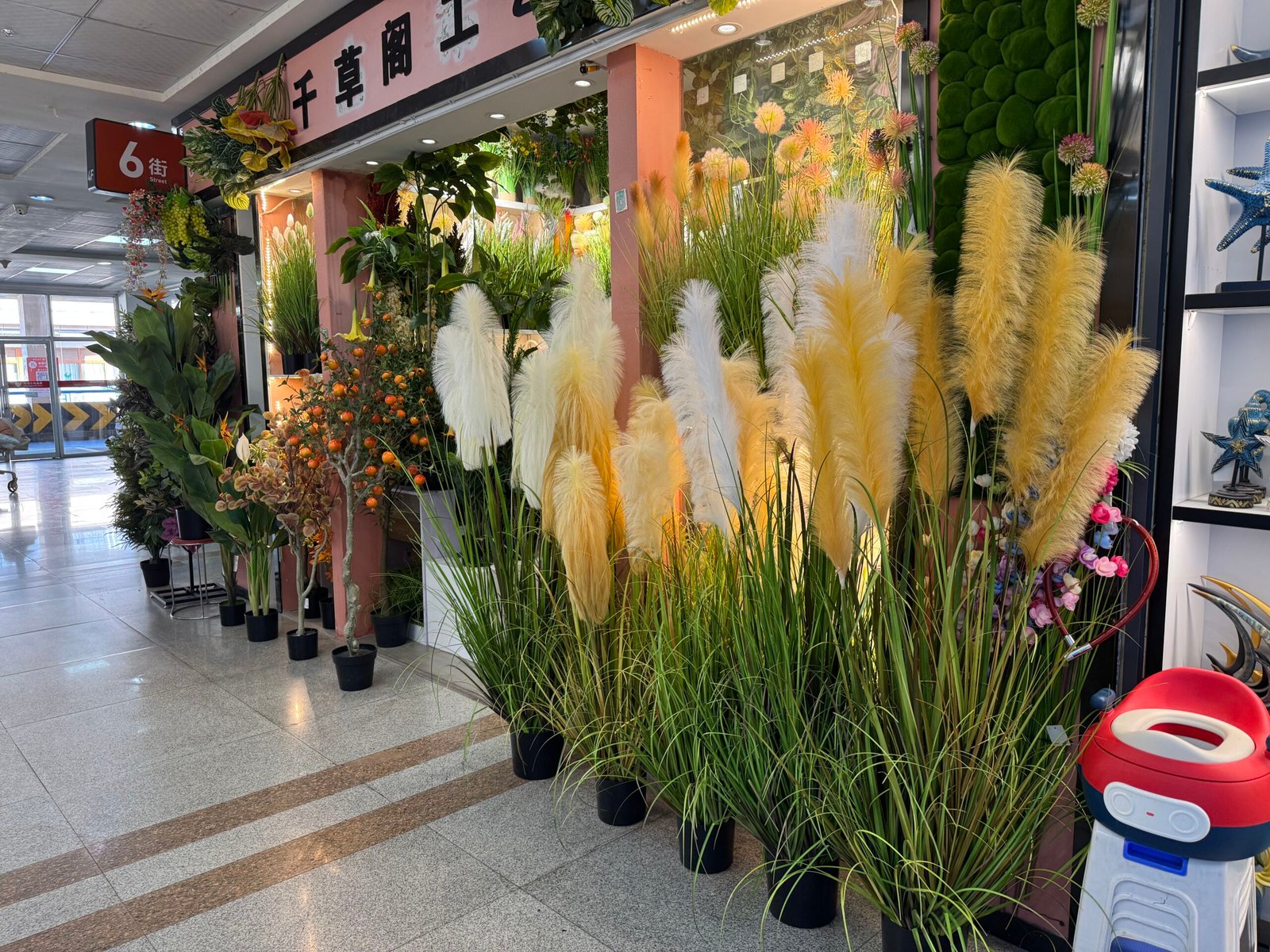 Yiwu home decor market storefront filled with tall yellow and white pampas grass, faux plants, and potted trees along the aisle.