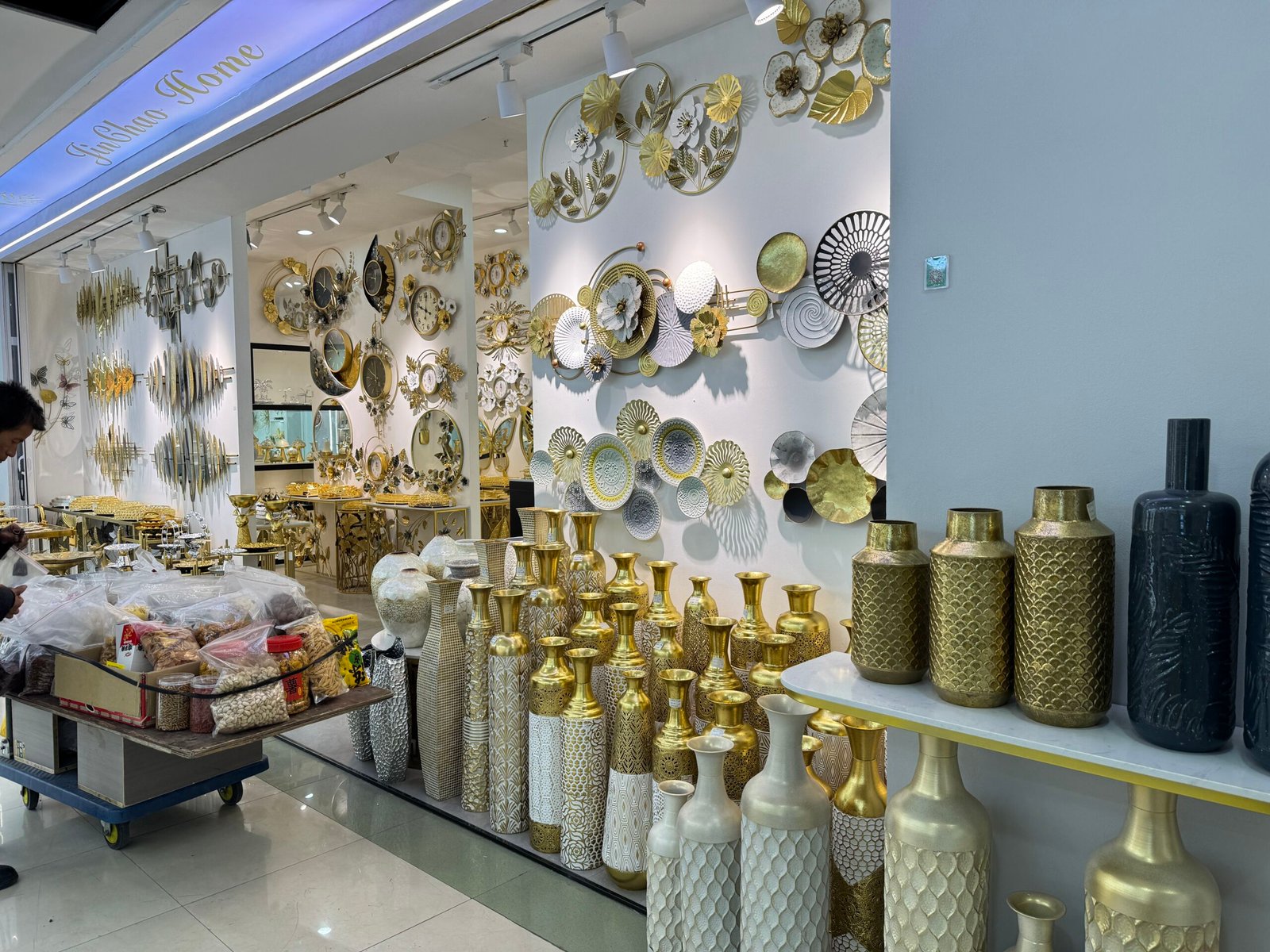 Fashion Home shop in Yiwu home decor market with its entrance lined by gold and white metal vases and interior walls covered in decorative metal wall art.