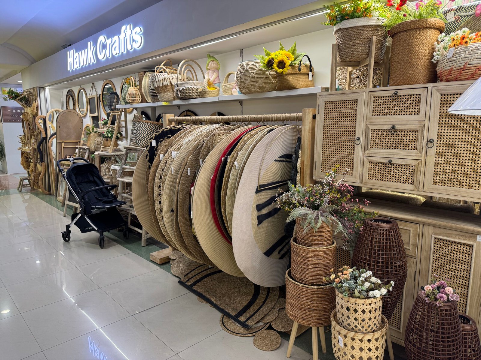 Hawk Crafts shop in Yiwu home decor market with stacks of round jute rugs, woven rattan baskets, cane-front cabinets, and wicker planters.