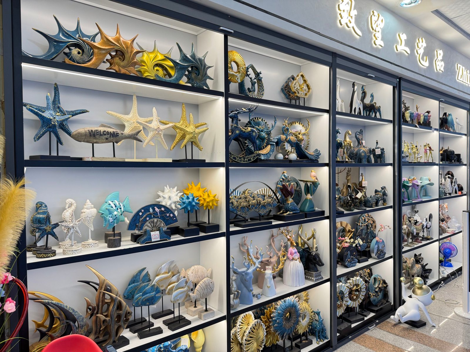 Yiwu decor shop shelves packed with resin starfish, abstract sea forms, fish figures, and fantasy sculptures in blue, gold, and neutral tones.