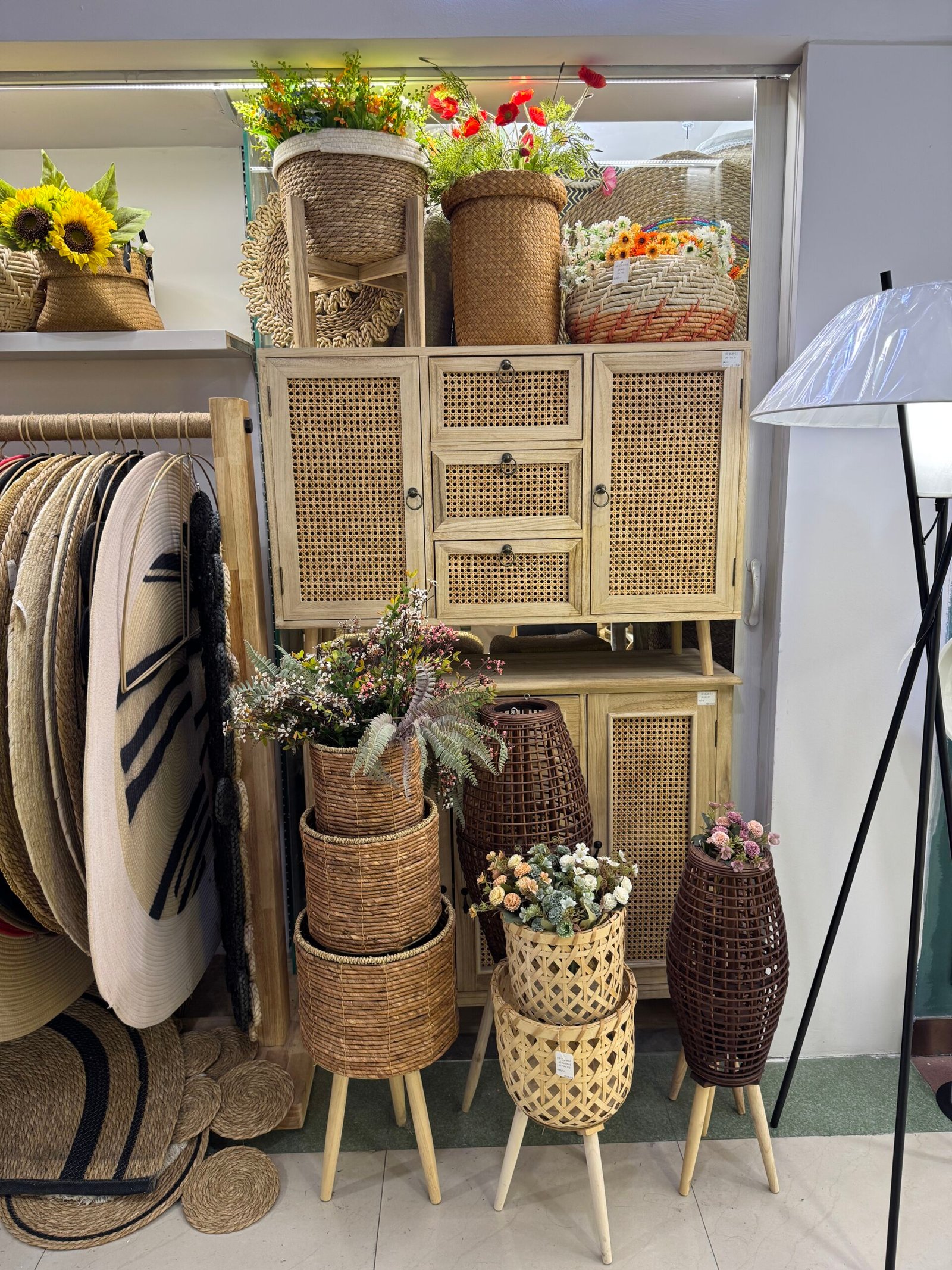 Yiwu home decor stall with a cane-front wooden cabinet, rattan planter stands, woven baskets, and colorful artificial flowers.